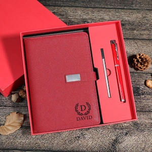 Puede incluir: Un juego de regalo rojo que incluye un cuaderno, un bolígrafo y un lápiz óptico. El cuaderno tiene un cierre plateado y el nombre "DAVID" con una inicial "D" y un diseño de corona de laurel. El juego se presenta en una caja roja.