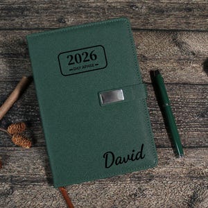 Puede incluir: Agenda verde de cuero de 2026 con cierre plateado y el nombre "David" grabado en la portada. Un bol&iacute;grafo verde a juego descansa junto a la agenda. La agenda est&aacute; sobre una superficie de madera.