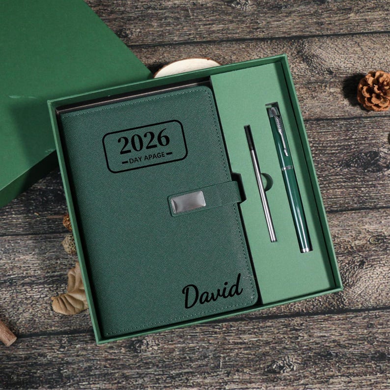Puede incluir: Un conjunto de regalo verde que incluye una agenda de cuero para 2026 con la inscripci&oacute;n "Day Apage", un bol&iacute;grafo y un l&aacute;piz &oacute;ptico. El nombre "David" est&aacute; impreso en la agenda.