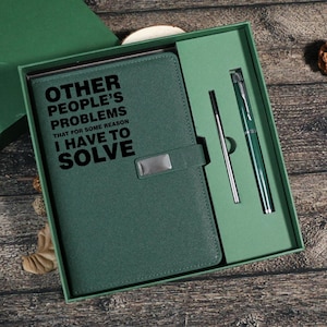 May include: A green notebook and pen set presented in a gift box. The notebook features the text "OTHER PEOPLE'S PROBLEMS THAT FOR SOME REASON I HAVE TO SOLVE". The pen is green with silver detailing. The set is in a green box.