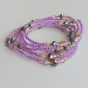 May include: A collection of purple beaded waist beads. The beads are a mix of small, round beads and larger, iridescent crystal beads. Gold-coloured spacers are interspersed throughout the strands. The waist beads are arranged in a circular pattern.