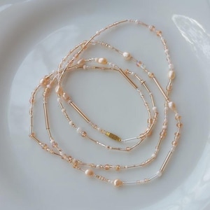 May include: Delicate body chain featuring a mix of small beads, pearls, and gold-colored metal accents. The chain is arranged in a flowing, layered design, with a subtle, elegant aesthetic. The beads are in shades of peach and cream.