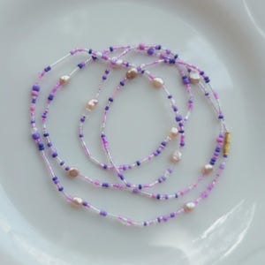 May include: A delicate necklace featuring a mix of small, colorful beads in shades of purple, pink, and white. The necklace also includes several irregularly shaped, light-colored pearls. The clasp is gold-colored.