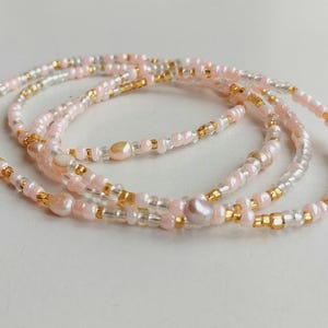 May include: Delicate beaded jewelry featuring strands of pink, gold, and clear beads. The strands are intertwined, creating a layered effect. The beads are small and round, with a mix of colors and textures. The jewelry is likely a waist bead or bracelet.