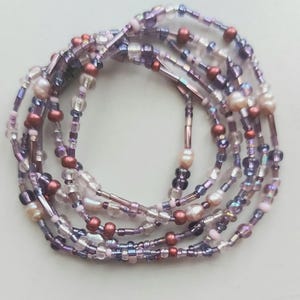 May include: A multi-strand beaded necklace in shades of purple, pink, and clear. The necklace features a variety of beads, including small seed beads, larger round beads, and some with a pearlescent finish. The strands are intertwined.