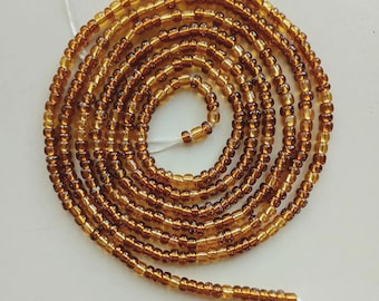 May include: A coiled strand of amber-colored waist beads. The beads are small, round, and have a glossy finish. The string is white and visible at the ends of the strand. The beads are arranged in a spiral pattern against a white background.