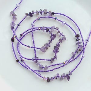 Puede incluir: Una colección de collares y cinturones de cuentas moradas. Los collares están hechos con pequeñas cuentas moradas claras y presentan acentos irregulares de piedra amatista. Algunas cuentas tienen un brillo iridiscente. Los artículos están dispuestos en espiral sobre una superficie blanca.