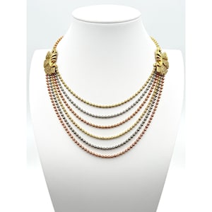 Puede incluir: Un collar de múltiples hilos con seis cadenas en cascada de pequeñas cuentas metálicas en tonos alternos de oro, plata y cobre. Los hilos están conectados a cada lado por un adorno de metal dorado texturizado en forma de abanico.