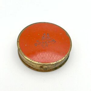 May include: A round, vintage compact with a bright orange lid and a gold-colored metal base. The lid features the word "Finesse" in cursive script, along with a small illustration. The compact is likely from the mid-20th century.