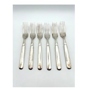 Puede incluir: Un juego de seis tenedores de metal plateado dispuestos en fila. Cada tenedor presenta cuatro dientes y un mango liso y recto con un pequeño detalle de monograma grabado cerca de la base. El metal tiene un acabado pulido y reflectante.