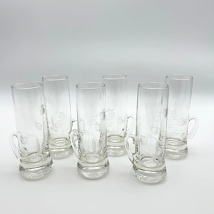 May include: Set of six clear glass shot glasses with handles. Each glass features an etched floral design. The glasses have a cylindrical shape with a slightly flared base. The handles are small and curved.