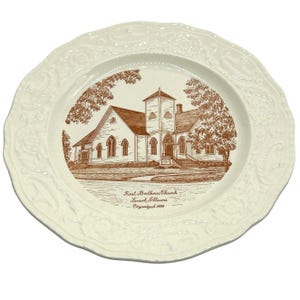May include: Off-white decorative plate featuring a sepia-toned illustration of the First Brethren Church in Lanark, Illinois, organized in 1884. The plate has a scalloped edge with embossed floral detailing.
