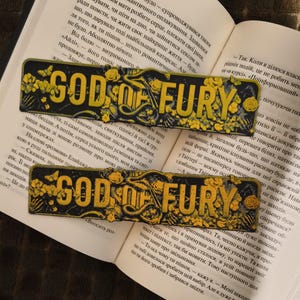 May include: Two black and yellow bookmarks, each with the text "GOD OF FURY", rest on an open book. The bookmarks have a distressed appearance, with floral and butterfly details.