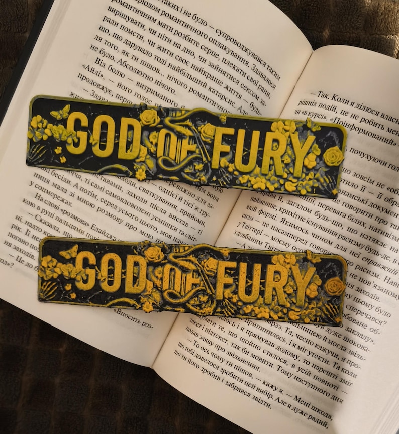 May include: Two black and yellow bookmarks, each with the text "GOD OF FURY", rest on an open book. The bookmarks have a distressed appearance, with floral and butterfly details.