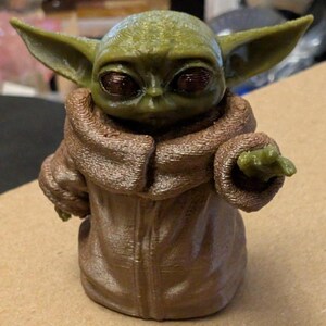 Puede incluir: Una peque&ntilde;a figura impresa en 3D de una criatura verde con grandes ojos oscuros y orejas puntiagudas. La figura viste una t&uacute;nica marr&oacute;n texturizada con cuello alto y tiene un brazo extendido hacia adelante con la mano abierta y l&iacute;neas visibles.