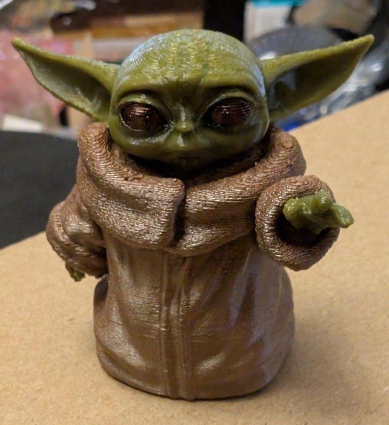 Puede incluir: Una peque&ntilde;a figura impresa en 3D de una criatura verde con grandes ojos oscuros y orejas puntiagudas. La figura viste una t&uacute;nica marr&oacute;n texturizada con cuello alto y tiene un brazo extendido hacia adelante con la mano abierta y l&iacute;neas visibles.