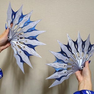 Puede incluir: Dos objetos decorativos en forma de abanico con un diseño en capas. Los abanicos presentan una combinación de colores plateado y azul con intrincados patrones geométricos. Los objetos se sostienen sobre un fondo neutro.