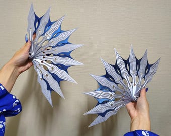 May include: Two decorative, fan-shaped objects with a layered design. The fans feature a silver and blue color scheme with intricate geometric patterns. The objects are held up against a neutral background.