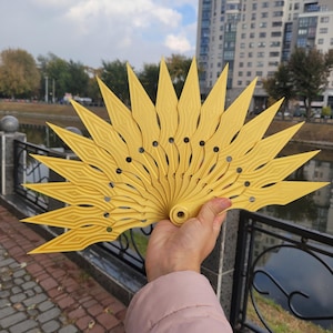 May include: A yellow fan with a sunburst design, held up against a backdrop of a cloudy sky and a building. The fan has pointed edges and small holes. The fan's design is reminiscent of a sun. The fan is made of a light material.