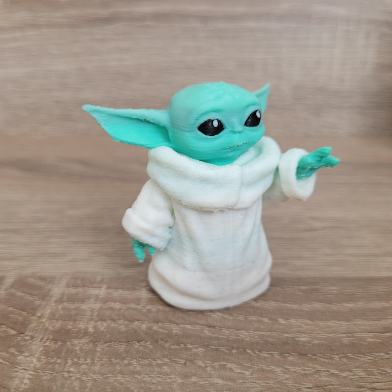 Puede incluir: Una peque&ntilde;a figura impresa en 3D de una criatura con piel de color verde menta claro, grandes orejas puntiagudas y ojos negros. La figura lleva una t&uacute;nica blanca texturizada con un cuello ancho y tiene una mano extendida hacia adelante.