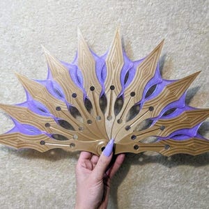 Könnte beinhalten: Ein handgeführter Faltfächer mit einer Reihe von miteinander verbundenen, spitz zulaufenden, goldfarbenen Lamellen. Zwischen den goldenen Lamellen befindet sich ein durchscheinendes, hellviolettes, netzartiges Material mit einem zentralen Drehpunkt.