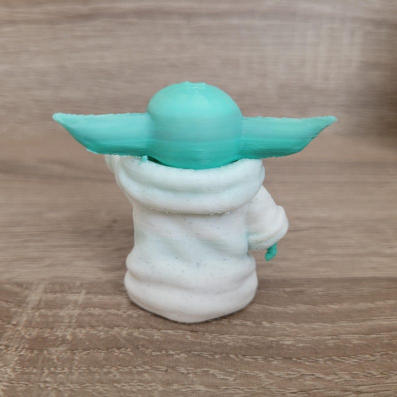 Puede incluir: Una peque&ntilde;a figura impresa en 3D que muestra una criatura con una cabeza lisa de color turquesa brillante y orejas grandes y puntiagudas. La criatura viste una t&uacute;nica texturizada de color blanco roto con capas horizontales visibles del proceso.