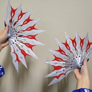 以下が含まれることがあります： 銀色と赤色の扇形の物体が2つあり、鋭く尖った刃が放射状に配置されています。各ピースには中央の回転軸があり、複数の細長いひし形のセグメントが重なり合って、装飾的で武器のようなデザインを形成しています。
