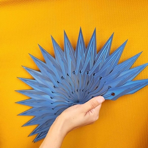 May include: A blue decorative fan with a radial design, held against a yellow background. The fan's blades have a layered appearance with subtle linear patterns. The fan is held in a hand, showcasing its size and shape.
