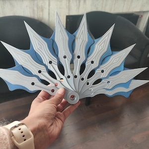 Puede incluir: Un abanico decorativo con un diseño en capas. El abanico presenta hojas plateadas con un patrón texturizado, acentuadas por bordes azules. El abanico es sostenido por una persona, mostrando sus intrincados detalles y artesanía.