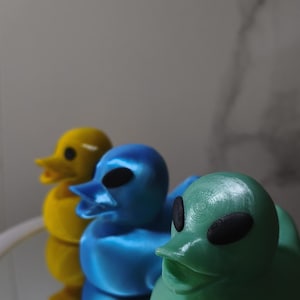May include: Three duck-shaped figurines with alien features, arranged in a row. The front figurine is light green, the middle is metallic blue, and the back is yellow. Each has large, black, oval-shaped eyes and a duck-like beak, with visible 3D print lines.