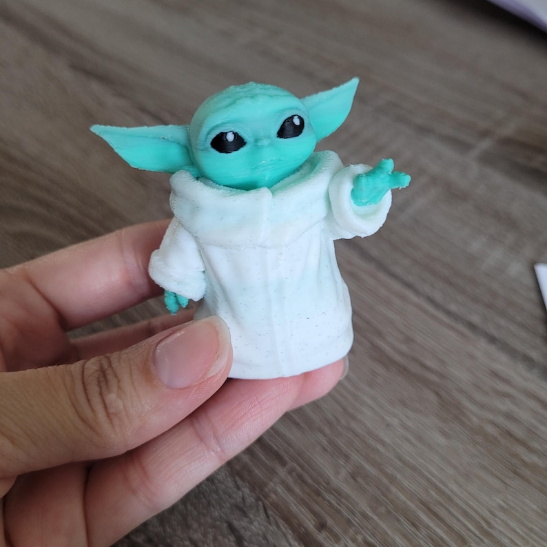 Puede incluir: Una peque&ntilde;a figura de color verde claro de una criatura con grandes orejas puntiagudas y ojos negros, que lleva una t&uacute;nica blanca texturizada con un cuello ancho. La figura se muestra con una mano levantada en un gesto y acabado mate.