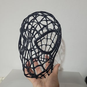 Puede incluir: Una máscara negra de tejido abierto que se asemeja a la cara de un superhéroe. La máscara presenta un diseño de telaraña y aberturas para los ojos. La máscara se sostiene en una mano sobre un fondo blanco. Es un artículo de cosplay o disfraz.