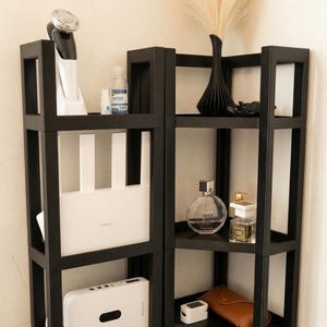 May include: A black modular corner shelving unit with three tiers, featuring an open frame design. The shelves hold various items including a white router, a white electronic device, a black vase with dried grass, perfume bottles, and a small brown case.