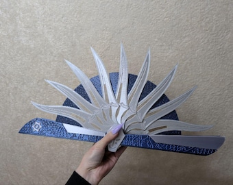May include: A handheld folding fan featuring a series of silver-colored blade-shaped slats arranged in a semi-circle. The base of the fan is a textured, deep blue material with a metallic finish and a small star-shaped emblem on the left side.
