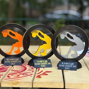 Może przedstawiać: Trzy trofea o tematyce koszykówki z czarnymi okrągłymi ramkami i sylwetkami graczy w kolorze pomarańczowym, żółtym i srebrnym. Każde trofeum ma czarną podstawę z tekstem "III MICLIE SKARB SUMMER JAM 1X1", "I MICLIE SKARB SUMMER JAM 1X1" i "II MICLIE SKARB SUMMER JAM 1X1".