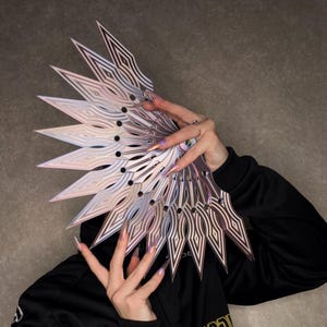 May include: A silver and iridescent geometric fan-shaped accessory with a layered design. The fan has a series of pointed, diamond-patterned blades. The accessory is held in front of a person, partially obscuring their face.