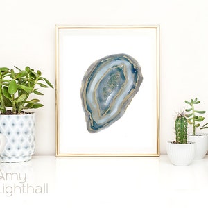 May include: A framed print featuring a watercolor depiction of a geode. The artwork showcases shades of blue, gray, and gold, with a gold frame. The print is displayed on a white surface, with potted plants in the foreground.