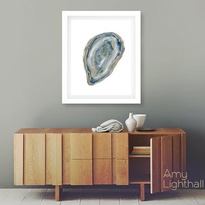 May include: Framed watercolor print of a blue and gold agate slice. The artwork is displayed in a white frame and hangs above a wooden cabinet. The cabinet has a natural wood finish and a modern design.