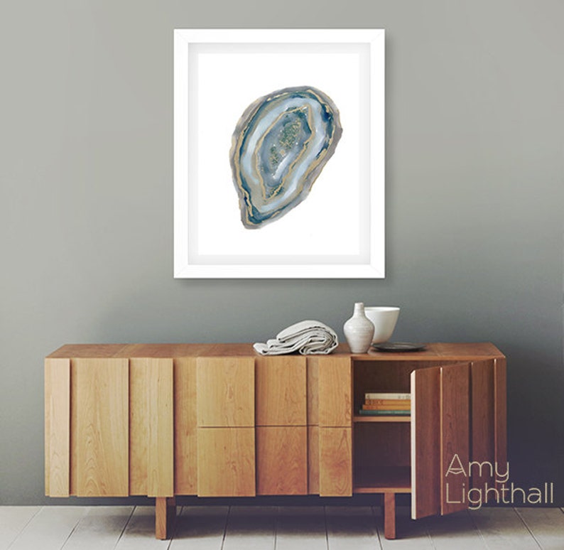 May include: Framed watercolor print of a blue and gold agate slice. The artwork is displayed in a white frame and hangs above a wooden cabinet. The cabinet has a natural wood finish and a modern design.