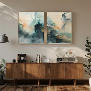 May include: Two rectangular wall art prints featuring abstract watercolor patterns in shades of deep teal, navy blue, and soft peach. The artwork is displayed in thin wooden frames above a long wooden sideboard cabinet with four cabinet doors.