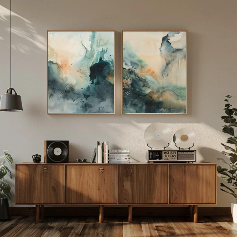 May include: Two rectangular wall art prints featuring abstract watercolor patterns in shades of deep teal, navy blue, and soft peach. The artwork is displayed in thin wooden frames above a long wooden sideboard cabinet with four cabinet doors.