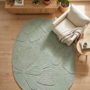 May include: Oval, light green rug with a floral pattern. The rug is placed on a wooden floor, with a cream-colored armchair and a small side table nearby. The rug's design features a large, stylized flower.