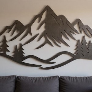 Op de afbeelding: Een donkerbruin houten wandkunstwerk dat een bergketen met dennenbomen afbeeldt. Het kunstwerk is bevestigd aan een lichtgekleurde muur boven een grijze bank. Het ontwerp heeft een golvende basislijn.