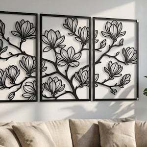 May include: A triptych of black metal wall art featuring a magnolia flower design. The artwork is composed of three rectangular panels, each framed in black, with the floral branches and blossoms intricately cut out. The panels are mounted on a white wall above a beige sofa.