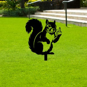 May include: Black metal squirrel garden stake. The silhouette depicts a squirrel giving the middle finger. The garden stake is inserted into the ground, and the squirrel is facing forward. The background is green grass.