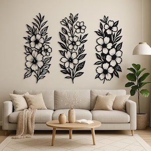 May include: Three black floral wall art pieces with intricate cut-out designs. Each piece features stylized flowers and leaves, mounted on a neutral-coloured wall above a sofa. The decor adds a touch of nature-inspired elegance to the room.