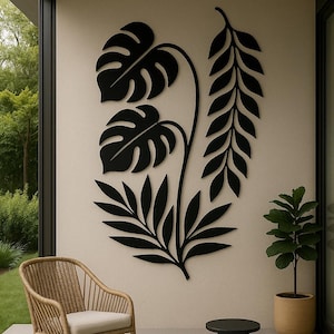 Puede incluir: Arte mural de metal negro con un diseño botánico estilizado de hojas y tallos. La obra de arte está montada en una pared de color neutro. Una silla de mimbre con un cojín crema y una pequeña mesa negra están en primer plano.