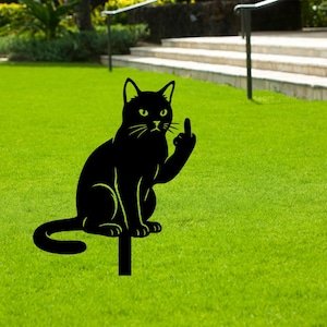 Peut inclure: Un piquet de jardin en silhouette de chat en métal noir. Le chat est assis et fait un doigt d'honneur. Le chat a des yeux jaunes et un contour jaune. Le piquet est dans un jardin herbeux.