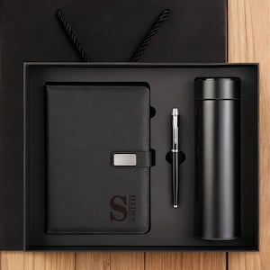 Puede incluir: Un juego de regalo negro que contiene un cuaderno, un bolígrafo y un vaso. El cuaderno tiene un cierre plateado y el nombre "SMITH" impreso. El bolígrafo y el vaso son negros. El juego se presenta en una caja negra y una bolsa de regalo negra.