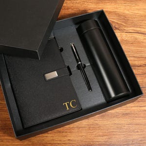 May include: A black gift set comprising a notebook, pen, and insulated flask, presented in a black box. The notebook features a silver clasp and gold lettering reading "TC". The flask and pen are black.